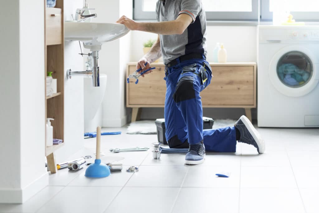 Plombier agenouillé répare un évier dans une salle de bain moderne. Outils et eau sur le sol carrelé. Machine à laver en arrière-plan.