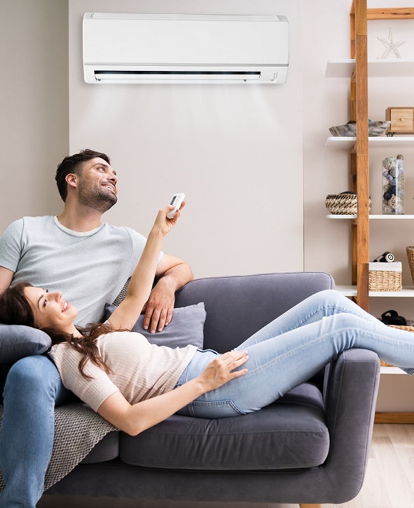 Couple souriant sur un canapé, réglant la climatisation avec une télécommande pour leur confort à la maison.